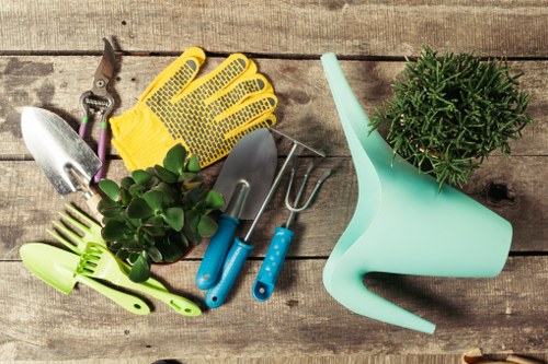 The image features a collection of gardening tools and accessories arranged on a weathered wooden surface, suitable for outdoor plant care and maintenance. Included are a pair of yellow gardening gloves with black rubber grip dots, a small silver hand trowel with a wooden handle, a silver transplanter with a blue handle, a green plastic plant water spray, and a pair of silver pruning scissors with purple handles. There is also a small potted plant with dense green foliage, possibly a herb or decorative shrub, positioned near the watering can. The tools and plant accessories are organized casually, emphasizing essential gardening equipment for tasks such as planting, watering, pruning, and soil planting. The overall setting suggests a well-prepared gardening workspace, ideal for residential gardens or outdoor landscaping projects typical in Crystal Palace and the surrounding areas, supporting professional gardening services like hedge trimming and garden maintenance offered by Hedge Trimming Crystal Palace.