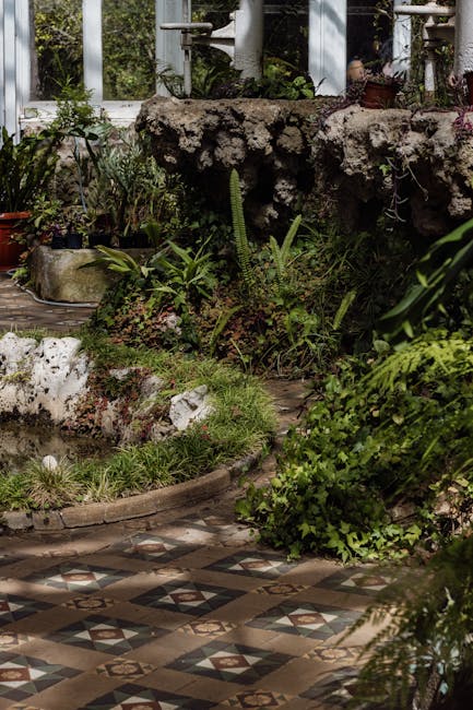 A well-maintained garden scene featuring a neatly trimmed hedge, lush green grass, and a variety of plants bordering a paved pathway. The hedge has been recently trimmed, displaying evenly cut foliage