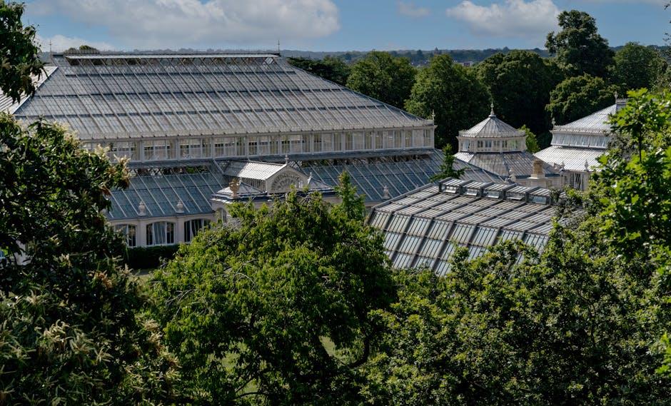 A large Victorian-style glasshouse with a metal framework, surrounded by lush green trees in a garden setting. The glasshouse features multiple sections with pitched roofs, some of which have decorati