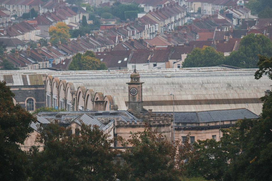 A panoramic view of a townscape with a prominent historic building featuring a clock tower and stone facade, surrounded by lush green trees and garden beds at the foreground. The garden area includes